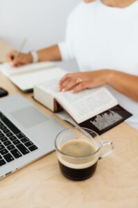 woman sitting at the desk with a laptop and a cup of coffee while using a paper dictionary